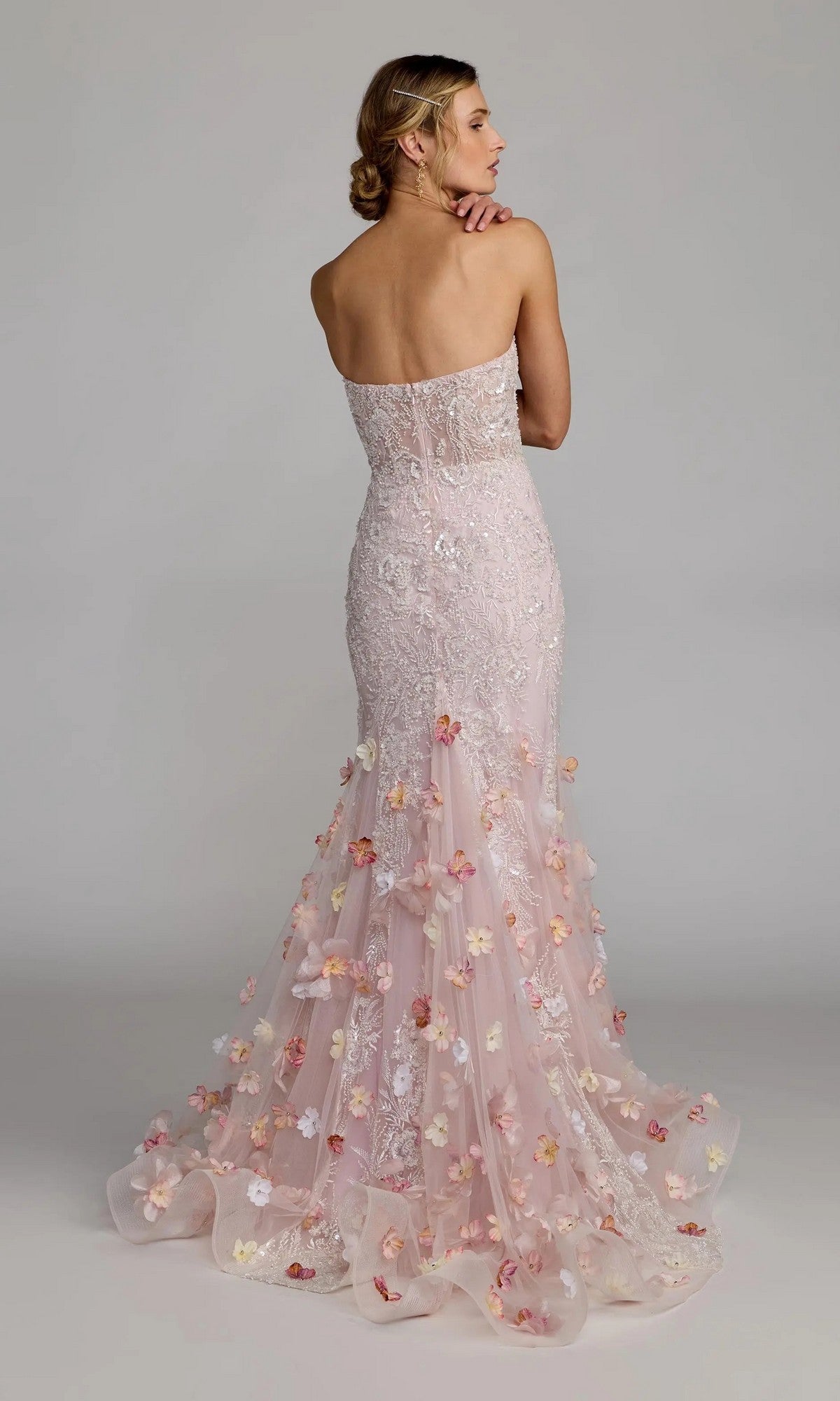 A woman stands facing away against a plain background, wearing the Long Formal Dress: Mac Duggal 21147—a strapless, pink mermaid gown with form-fitting silhouette and lace floral appliqués.